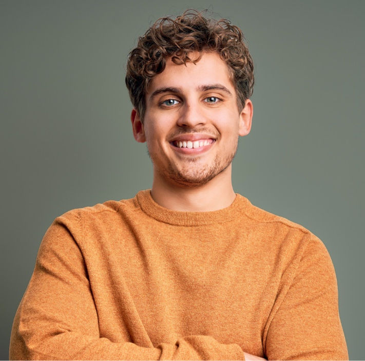 Man with short curly hair smiling with arms crossed, wearing a rust-colored sweater against a muted green background.