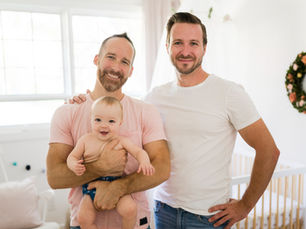 Two male parents holding their baby together in a calm, cozy nursery setting. Source: Getty Images. Credit: LSOphoto
