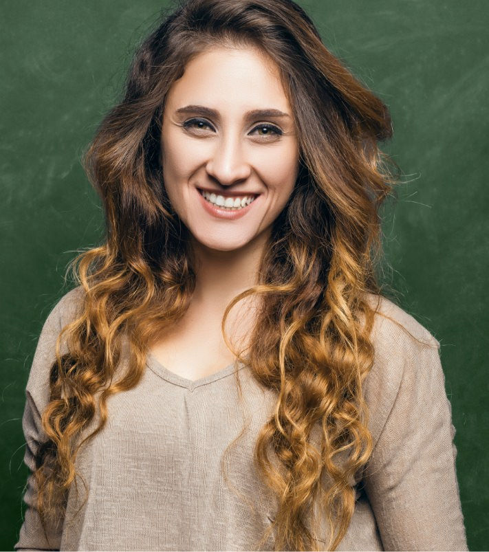 Woman with long wavy brown hair smiling, wearing a light beige top against a green background.