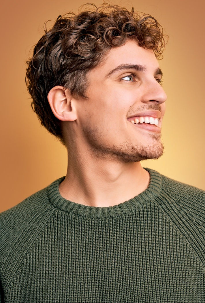 Man with short curly hair smiling and looking to the side, wearing a green knit sweater against a warm orange background.